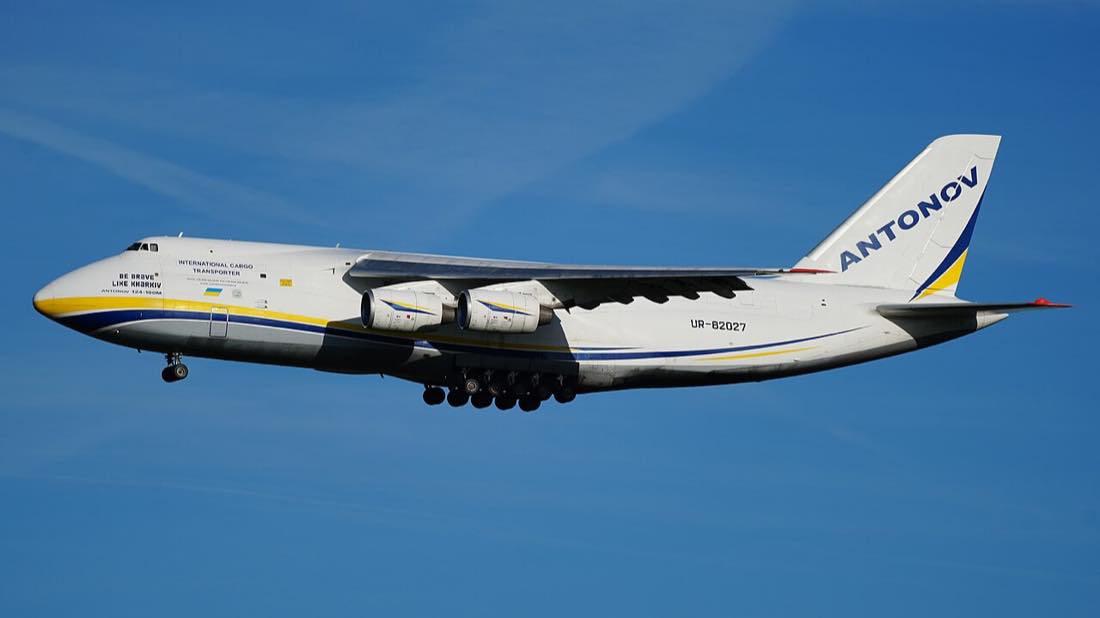 A white Antonov An-124 cargo plane in Ukrainian blue-and-yellow livery flying against a blue sky.